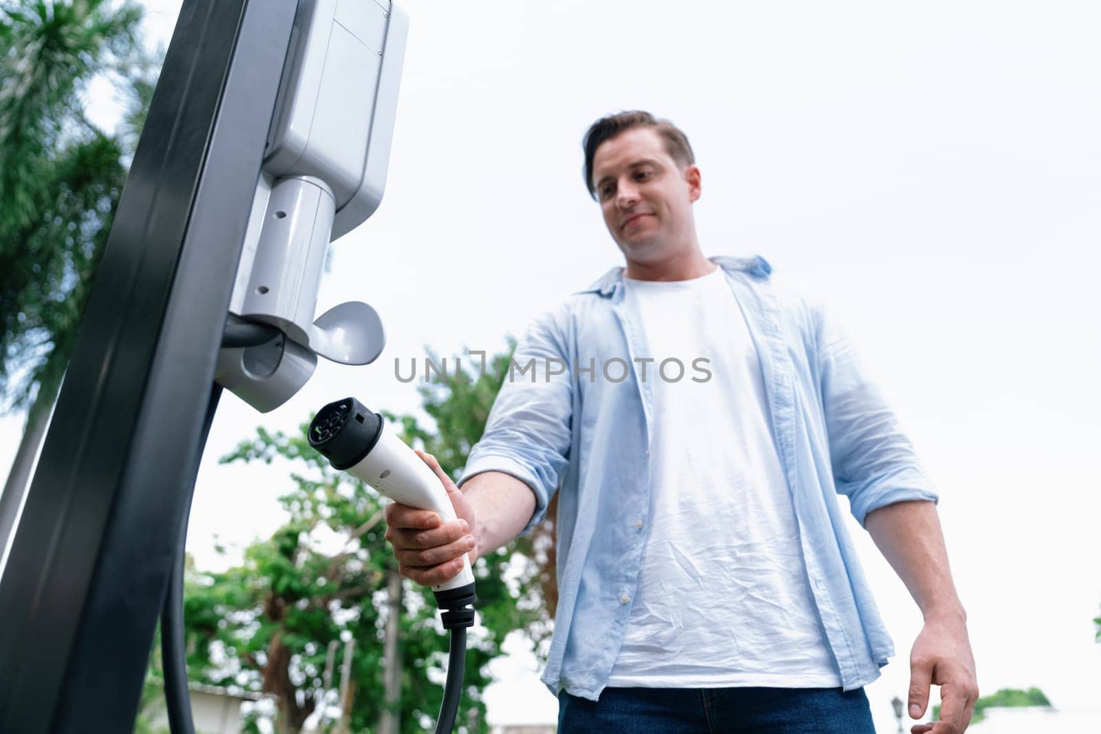 Eco-friendly conscious man recharging modern electric vehicle from EV charging station. EV car technology utilized as alternative transportation for future sustainability. Synchronos