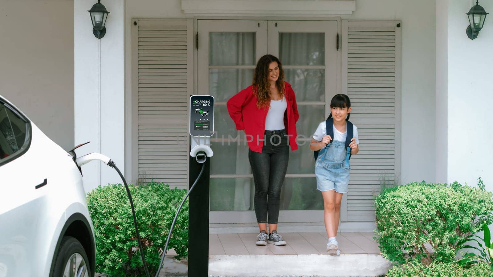 Happy little young girl learn about eco-friendly and energy sustainability as she help her mother recharge electric vehicle from home EV charging station. EV car and modern family. Panorama Synchronos