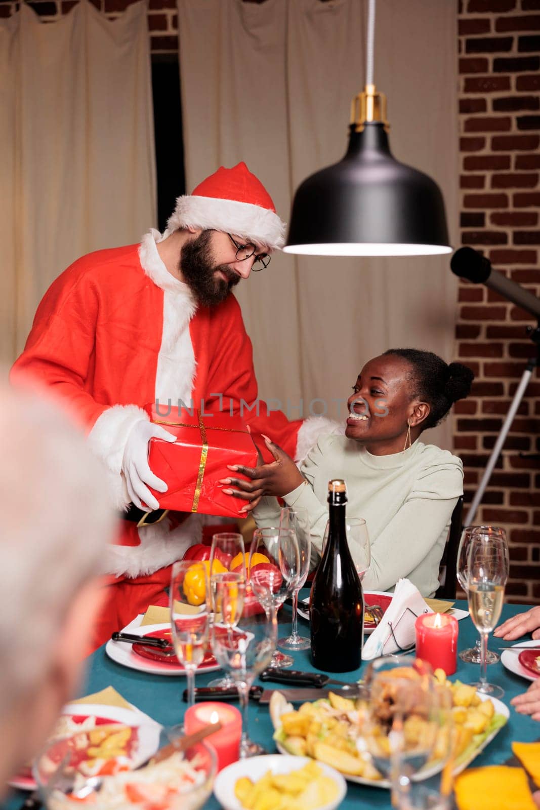 Santa claus gives present to african american woman celebrating christmas holiday with friends, xmas positivity and happiness. Person in red costume and hat feeling cheerful at home with family.