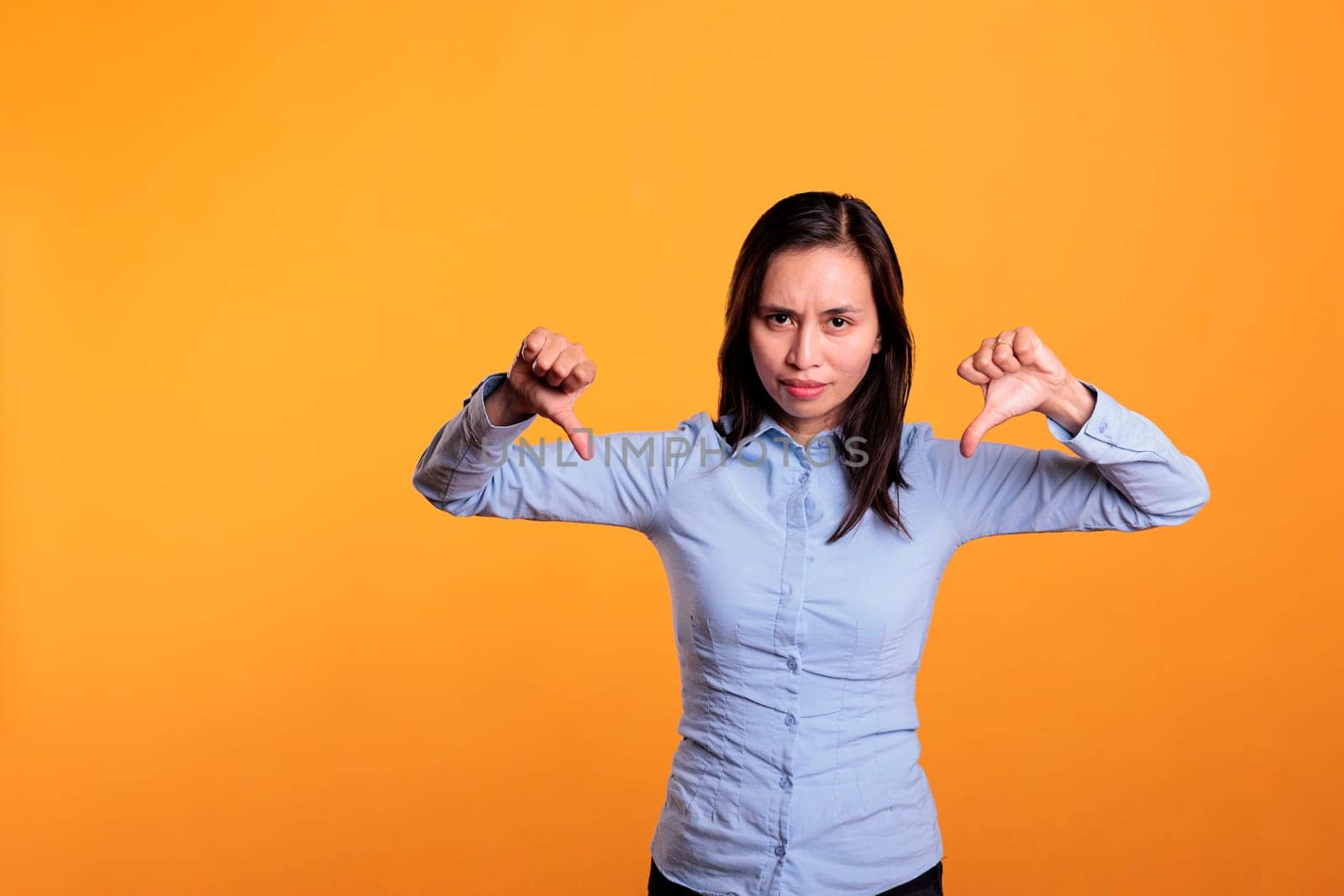 Unhappy filipino woman showing dislike gesture on camera in studio, gesturing disagreement sign with fingers. Displeased young adult expressing disapproval and disappointment sign. Dislike symbol