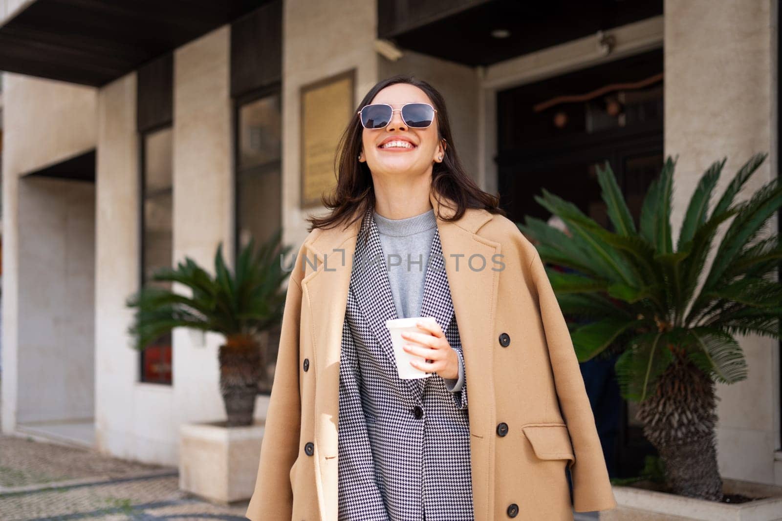 Happy smiling woman in sunglasses and coat walks out of hotel by andreonegin