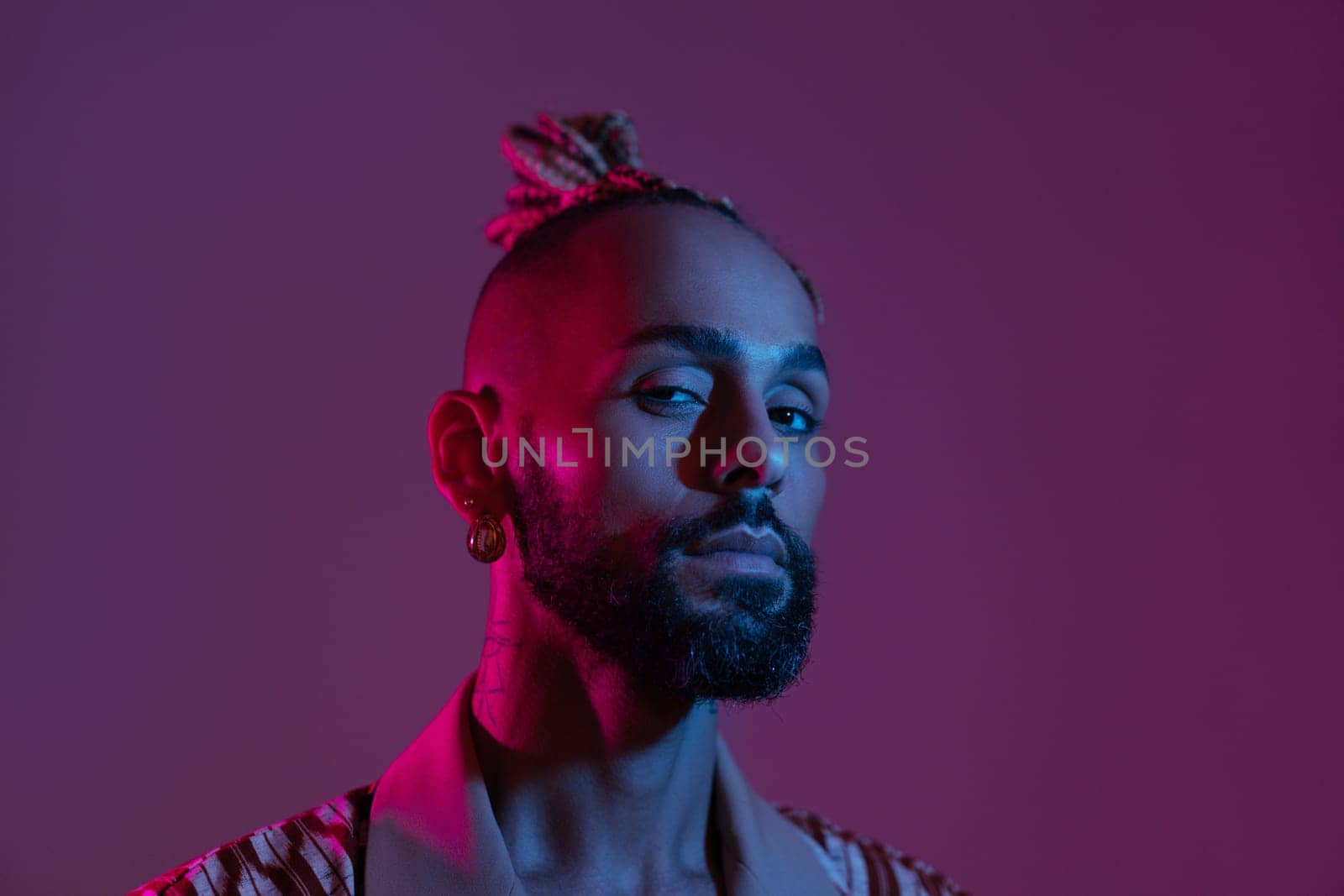 Latino gay man poses with bright makeup in neon light. Stylish man standing isolated background studio look in camera. Latino man with eyeshadows over standing looks at camera