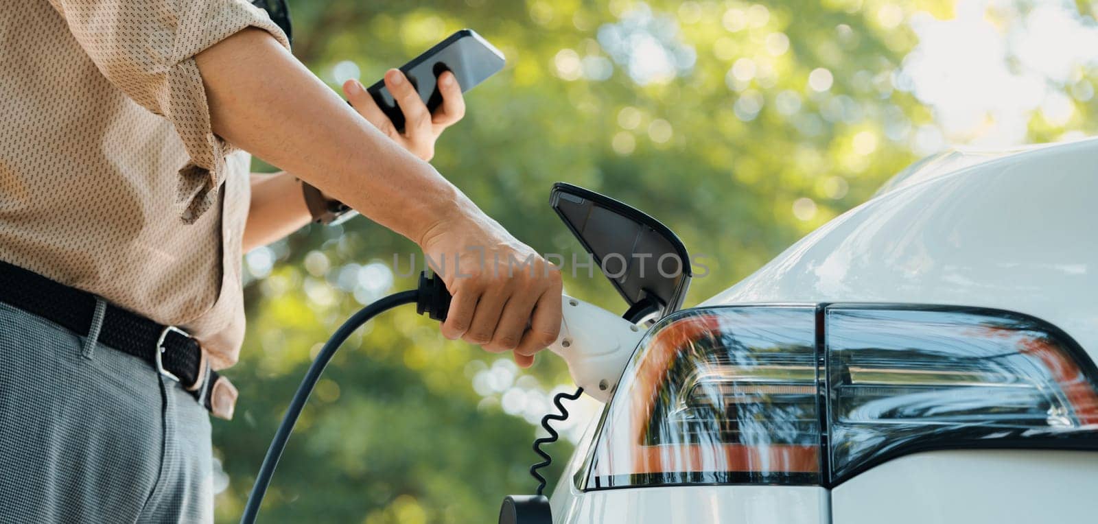 Young man use smartphone to pay for electricity at public EV car charging station green city park. Modern environmental and sustainable urban lifestyle with EV vehicle. Panorama Expedient