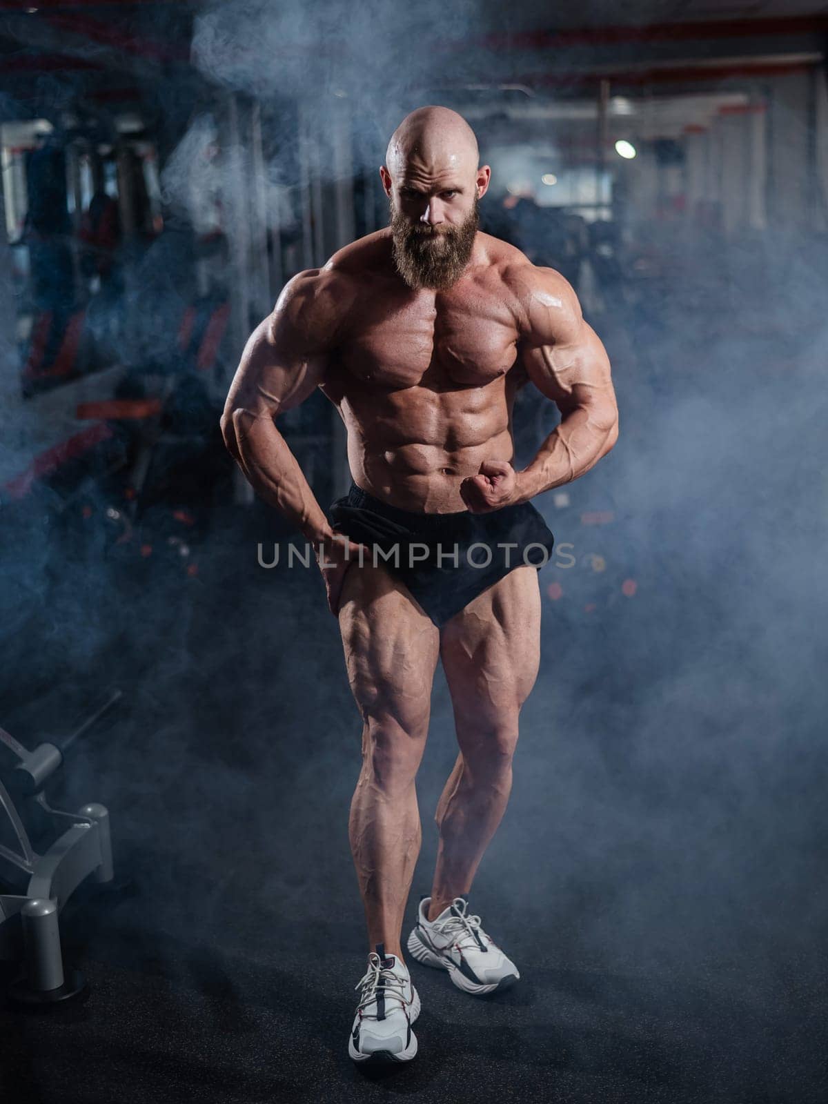 Muscular bald man posing in shorts. Bodybuilder showing off his shape in the gym