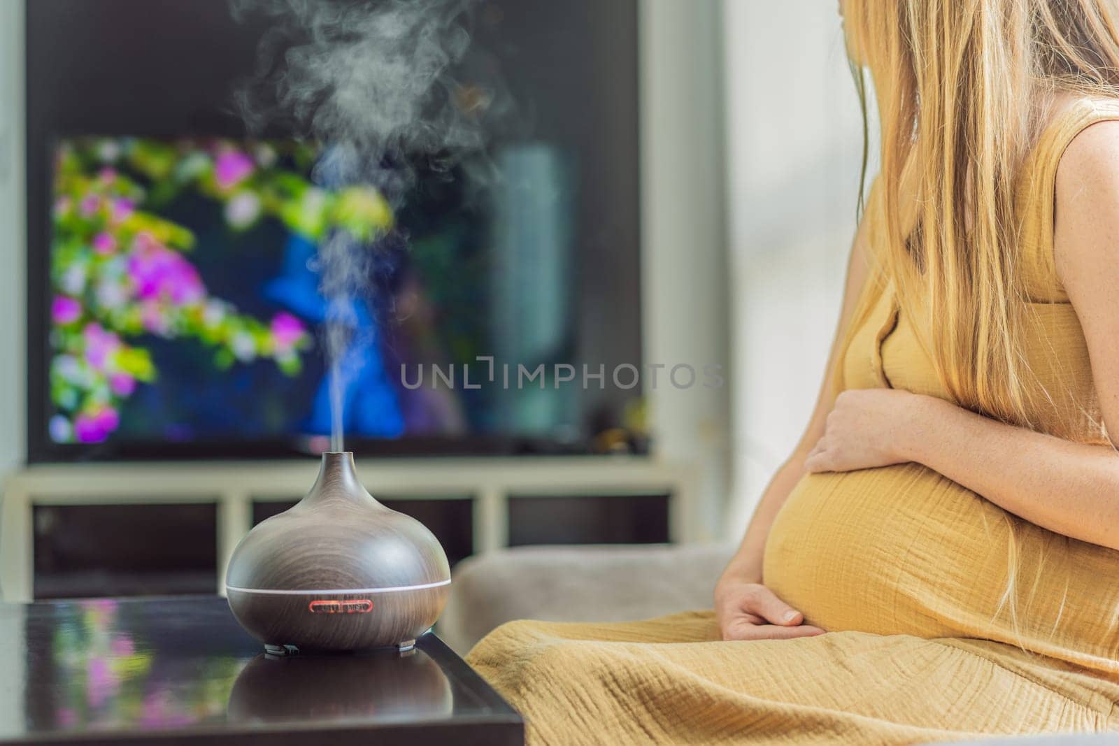 A blissful pregnant woman immerses in relaxation, savoring the soothing aroma from a diffuser while indulging in a calming TV video, embracing tranquility during her pregnant journey by galitskaya