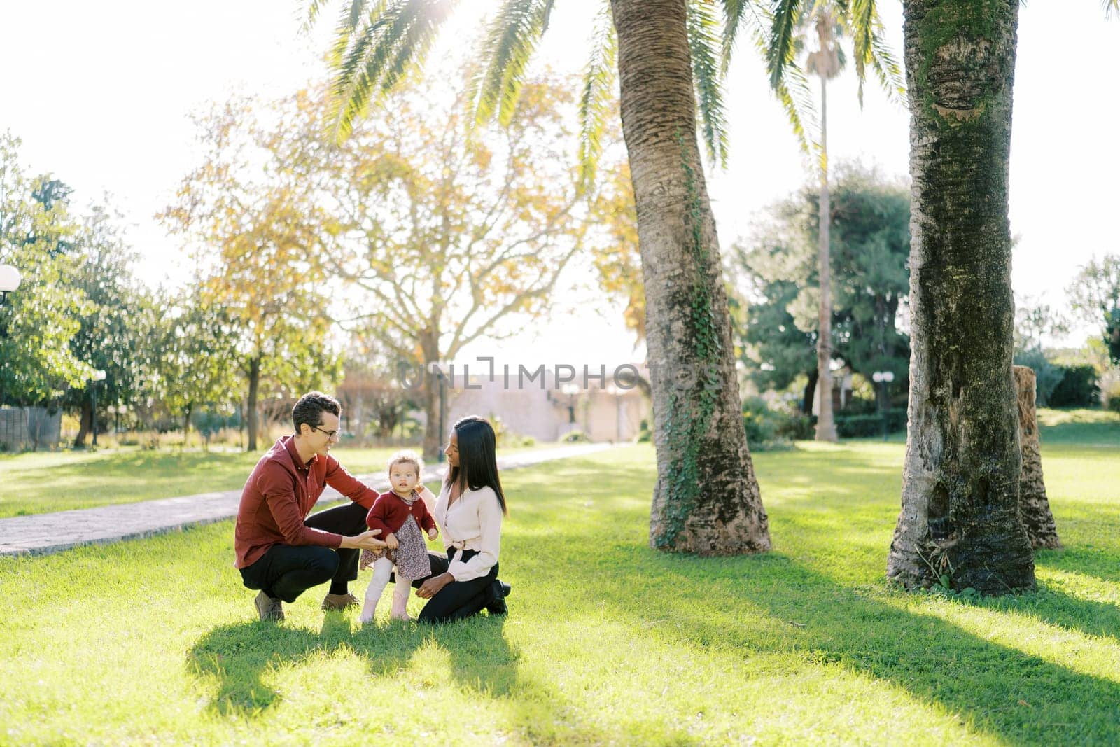 Parents squatting and looking at little girl in sunny park. High quality photo