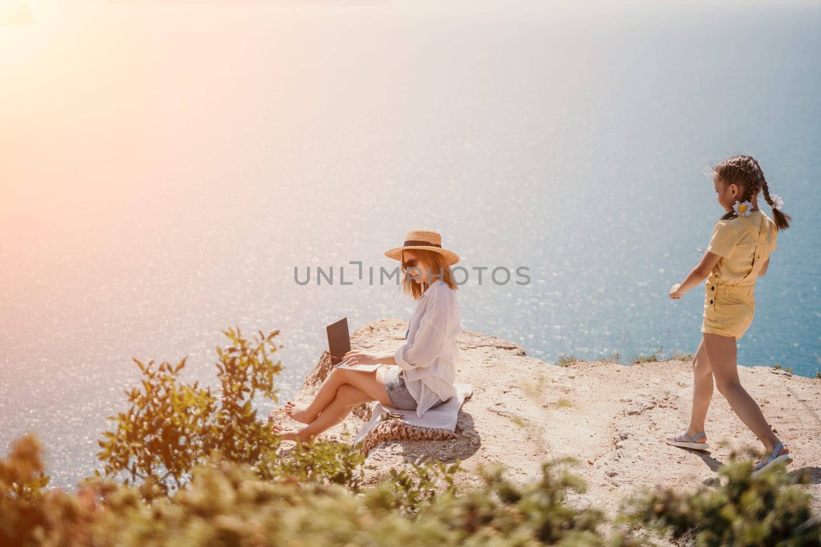 Successful business woman in yellow hat working on laptop by the sea. Pretty lady typing on computer at summer day outdoors. Freelance, travel and holidays concept.