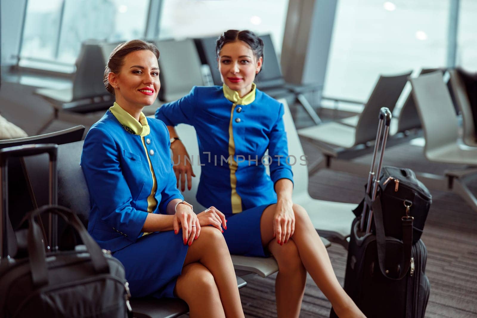 Female flight attendants in air hostess uniform looking at camera and smiling while sitting on chairs in passenger departure lounge