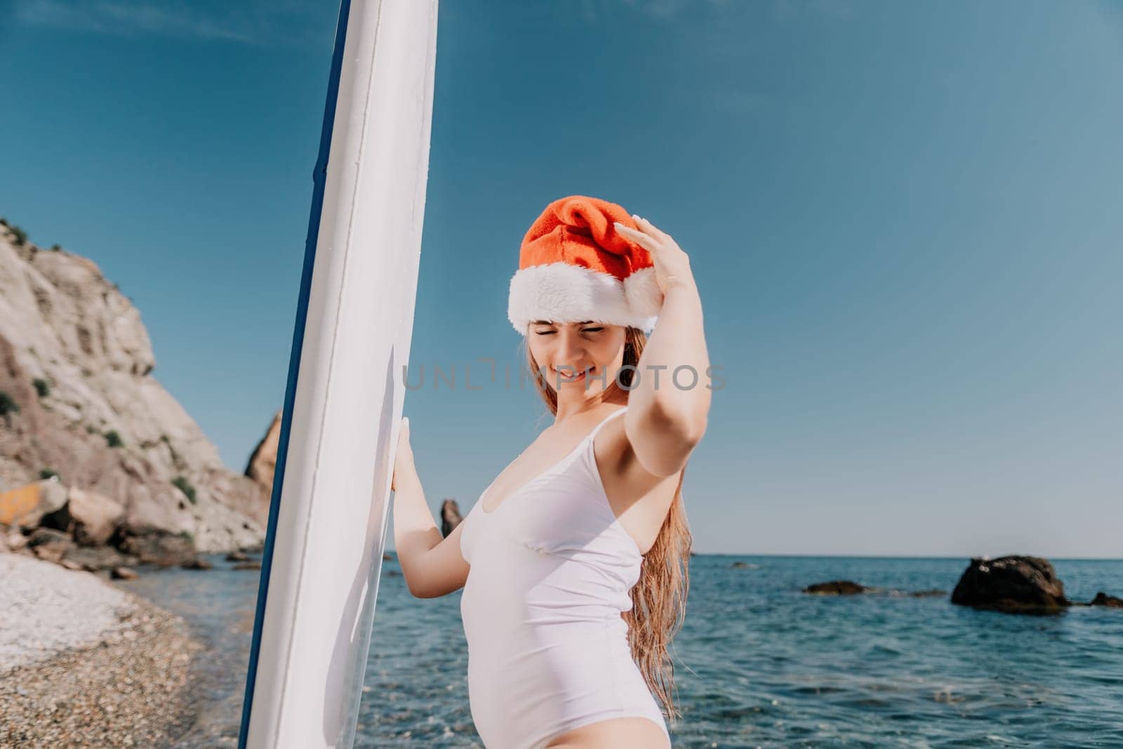 Woman sea sup. Close up portrait of happy young caucasian woman with long hair in Santa hat looking at camera and smiling. Cute woman portrait in a white bikini posing on sup board in the sea by panophotograph