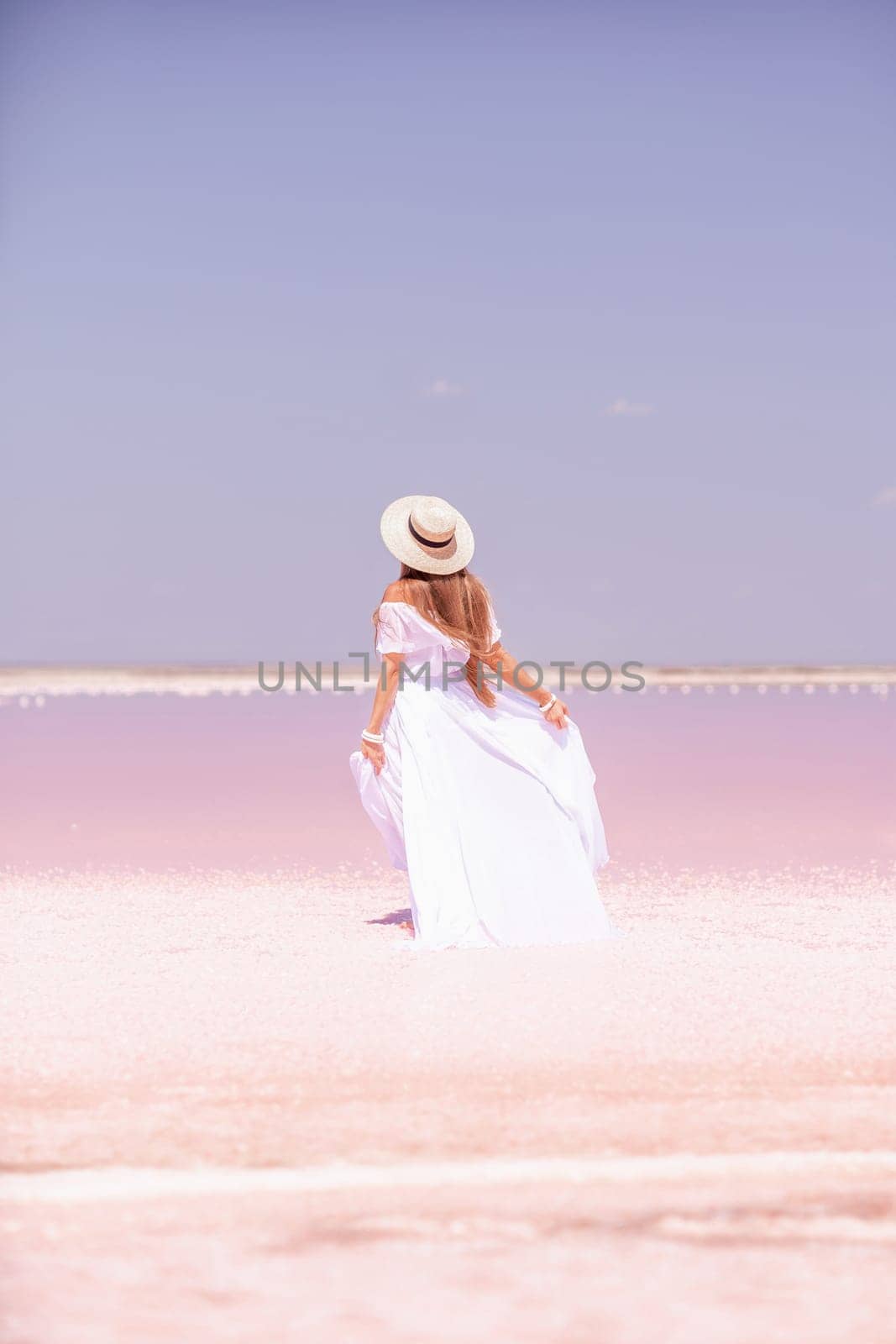 Woman in pink salt lake. She in a white dress and hat enjoys the scenic view of a pink salt lake as she walks along the white, salty shore, creating a lasting memory. by Matiunina