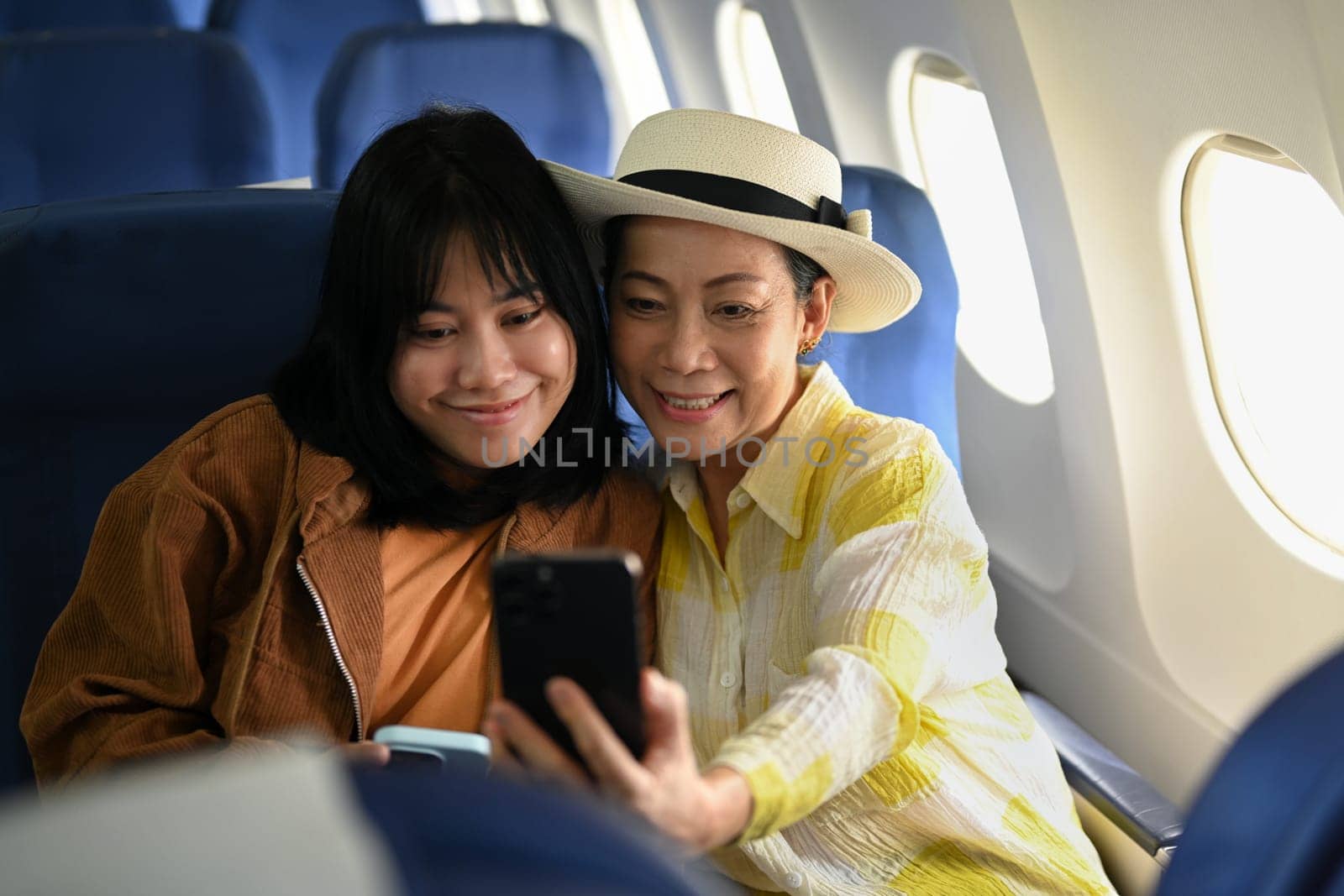 Happy 60s woman and daughter taking photo with smartphone in passenger airplane. Transportation, travel and summer vacation.