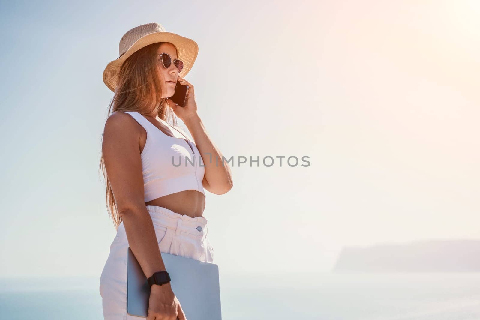Digital nomad, woman in the hat, a business woman with a laptop sits on the rocks by the sea during sunset, makes a business transaction online from a distance. Freelance, remote work on vacation. by panophotograph