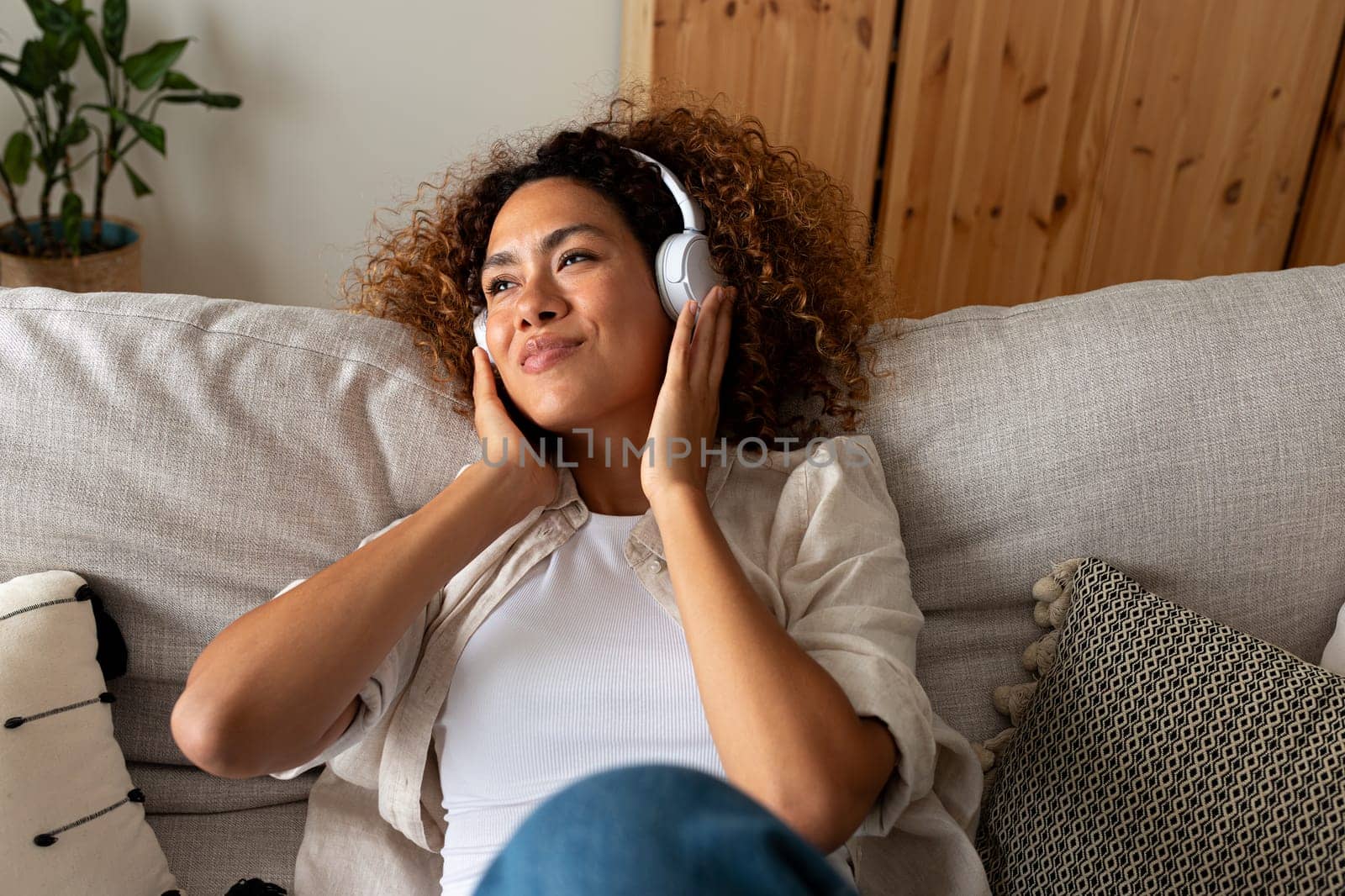 Relaxed young multiracial woman sitting on the sofa listening to music with wireless headphones. Lifestyle concept.