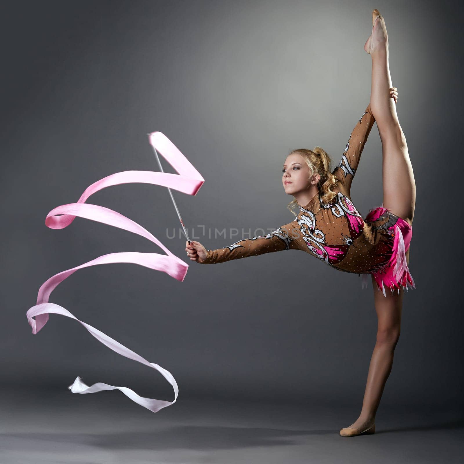 Rhythmic gymnast doing vertical split with ribbon, on grey background