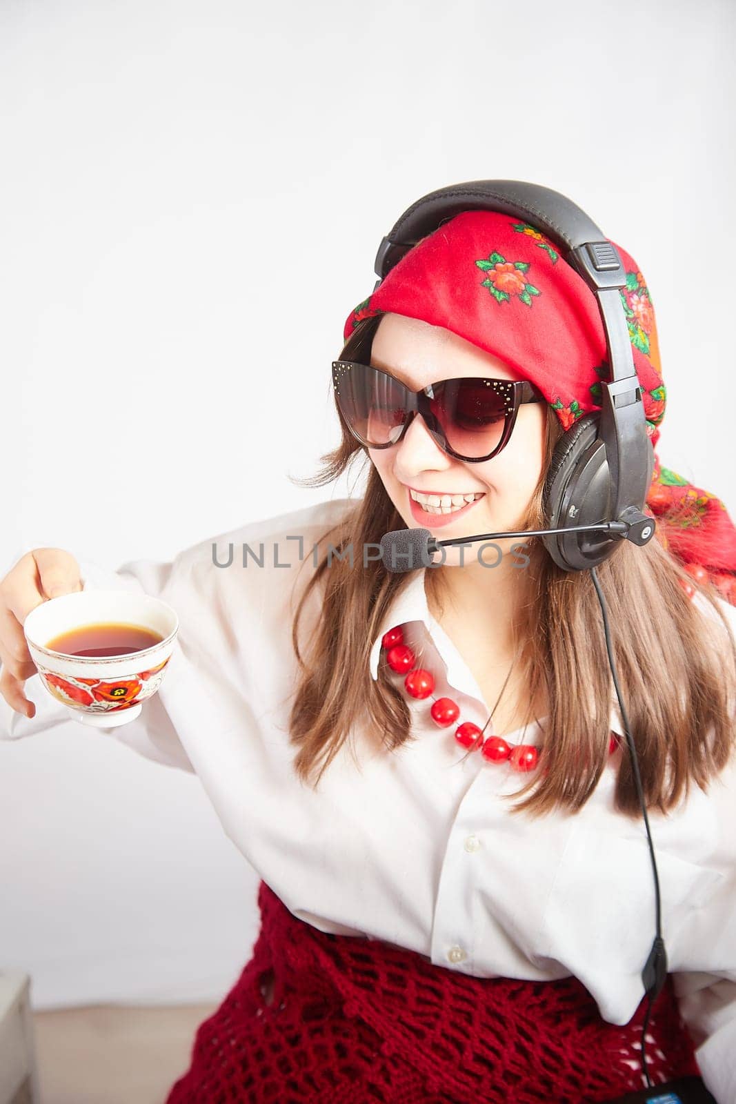 Portrait of young girl in a bright red scarf and large headphones with a microphone. A woman who is radio or television presenter with cap of tee. Funny female telecom operator. Freelancer at work