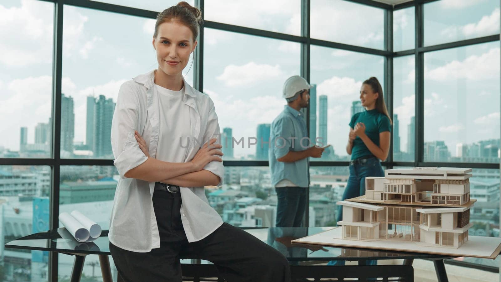 Young beautiful businesswoman sitting with arm folded while colleague discussion about house construction. Smart skilled architect engineer looking and smiling at camera with confident. Tracery