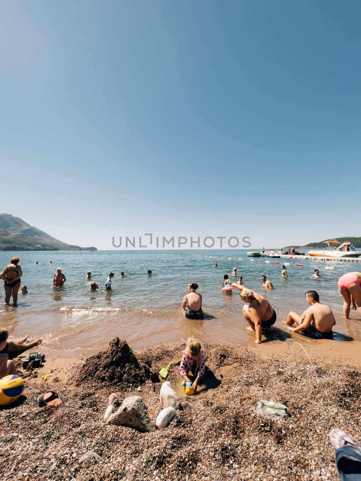 Little girl digs a hole in the sand on the beach against the backdrop of people bathing in the sea. High quality photo
