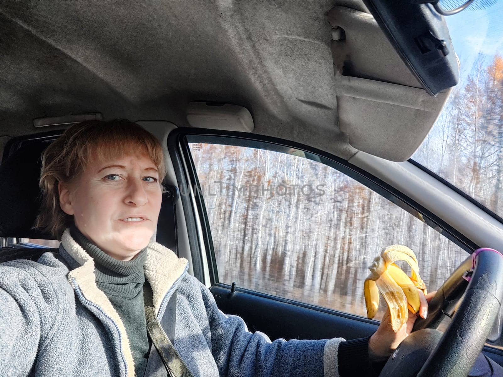 Portrait of female driver in solo journey. Adult mature middle aged woman holding steering wheel and a banana. Eating while driving for to stay awake. Lady girl with food while travel or trip by car
