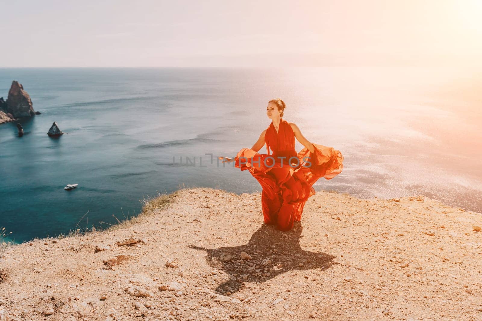 Side view a Young beautiful sensual woman in a red long dress posing on a rock high above the sea during sunrise. Girl on the nature on blue sky background. Fashion photo.