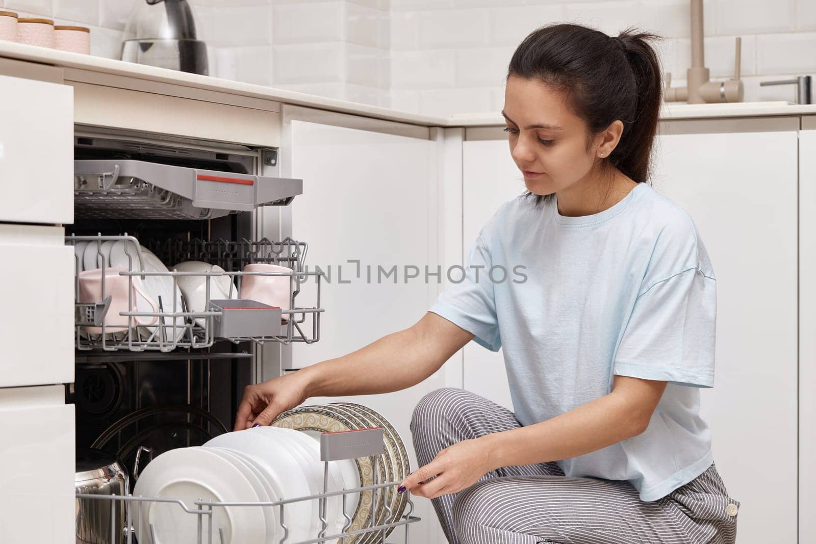 happy woman unloading plate from open automatic built-in dishwasher machine with clean utensils inside in modern white kitchen.