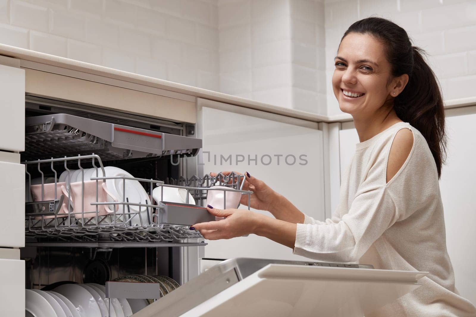 attractive woman unloading cup from open automatic built-in dishwasher machine with clean utensils inside in modern white kitchen.