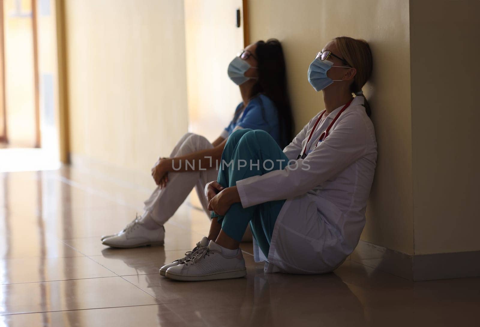 Young female doctor and nurse in medical masks sit in corridor of hospital. Emotional burnout of healthcare workers during covid19 pandemic concept.