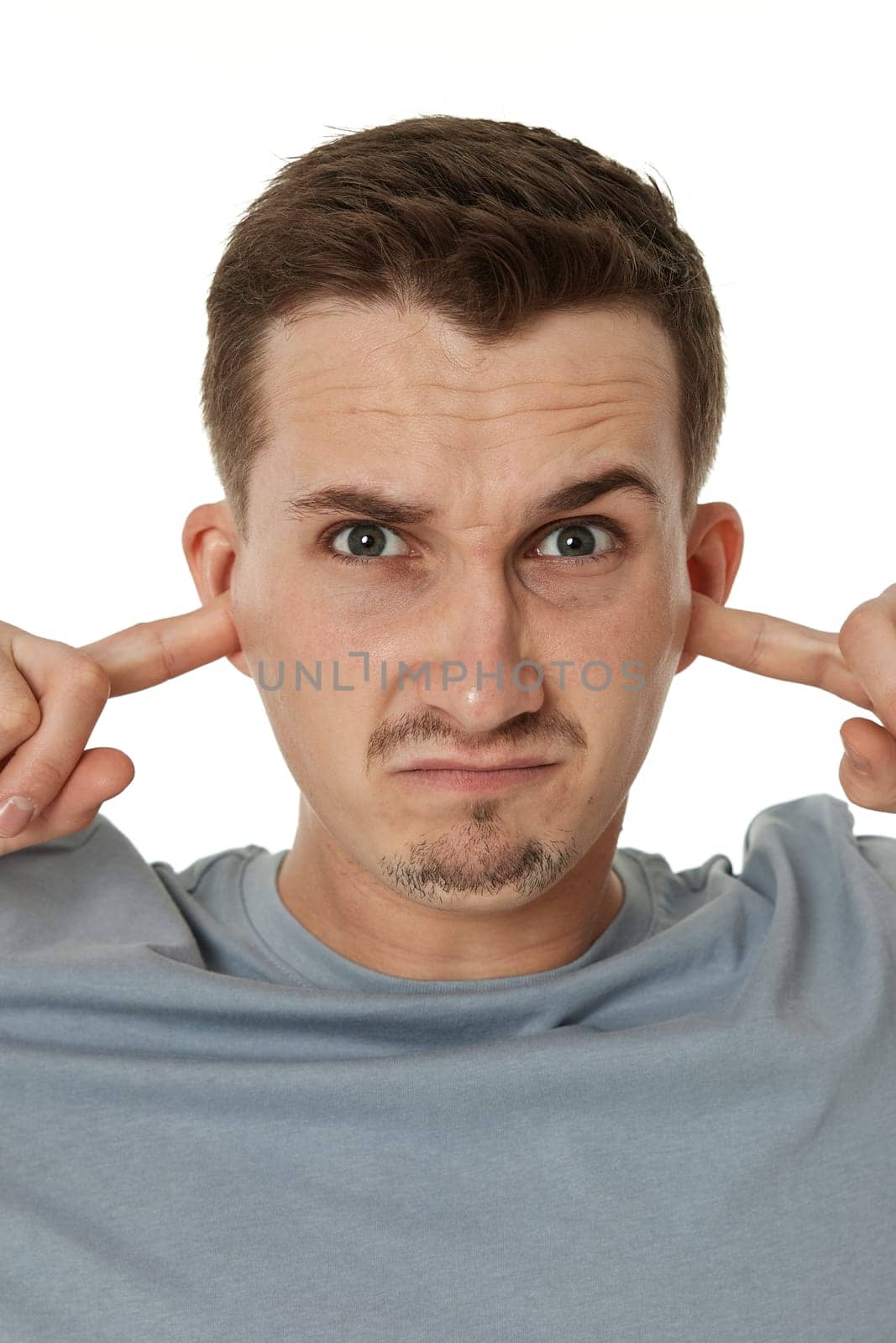 Young annoyed caucasian man in t-shirt covers ears with hands fingers on white background