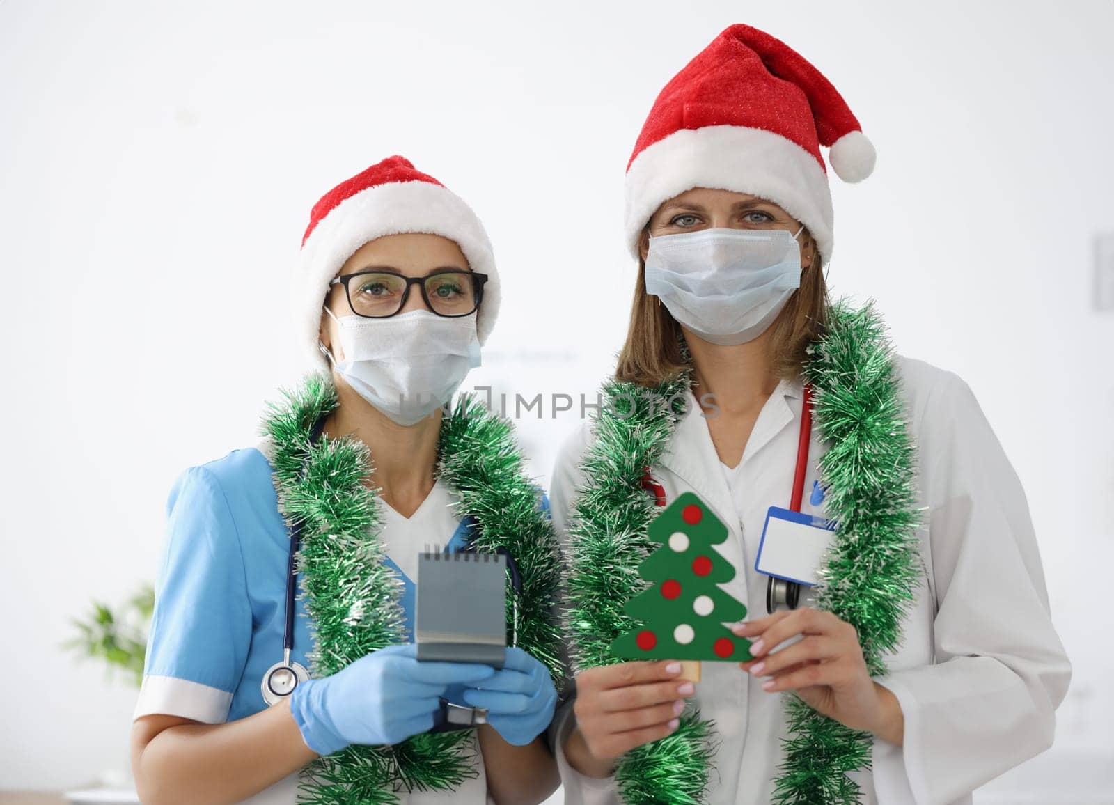 Nurse in blue suit, protective mask and gloves hold calendar with numbers 2021. Doctor in white coat hold paper Christmas tree with decorations in hand. Two women in santa claus hat and tinsel.