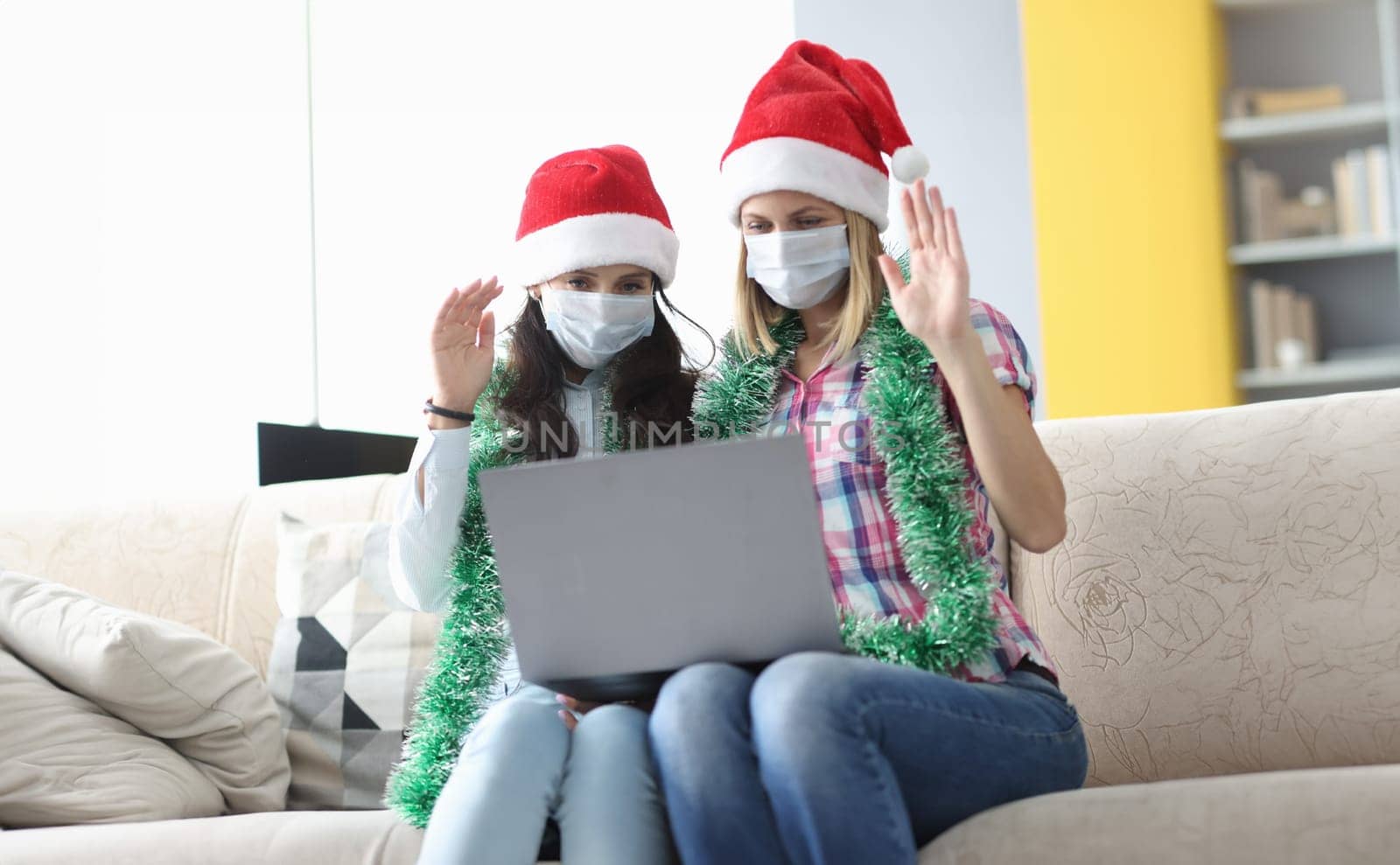 Two woman in protective mask and santa claus hat sit side by side and wave hello. Girlfriend talk via laptop on conference apps.