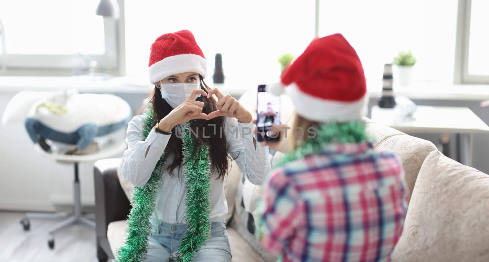 Woman in red hat and tinsel take photo on phone. Girlfriend in protective mask pose and make heart sign with hand.