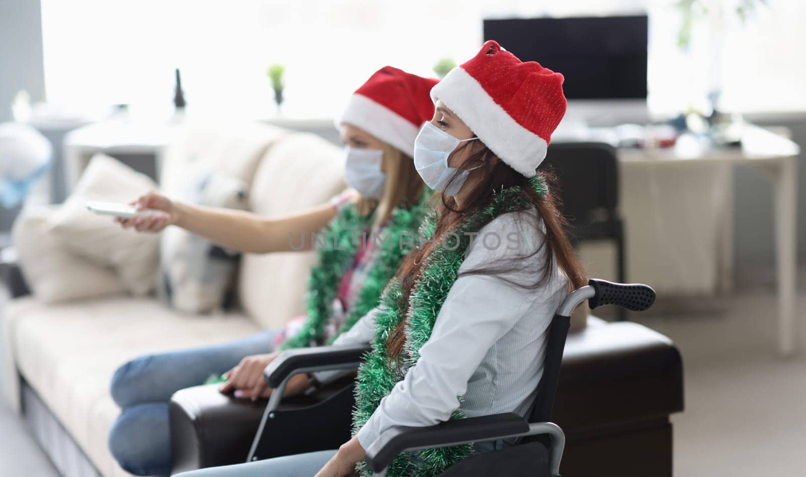 Woman in red cap and protective mask sit on couch and presses buttons on remote control. Friends sit in wheelchair and watch TV.