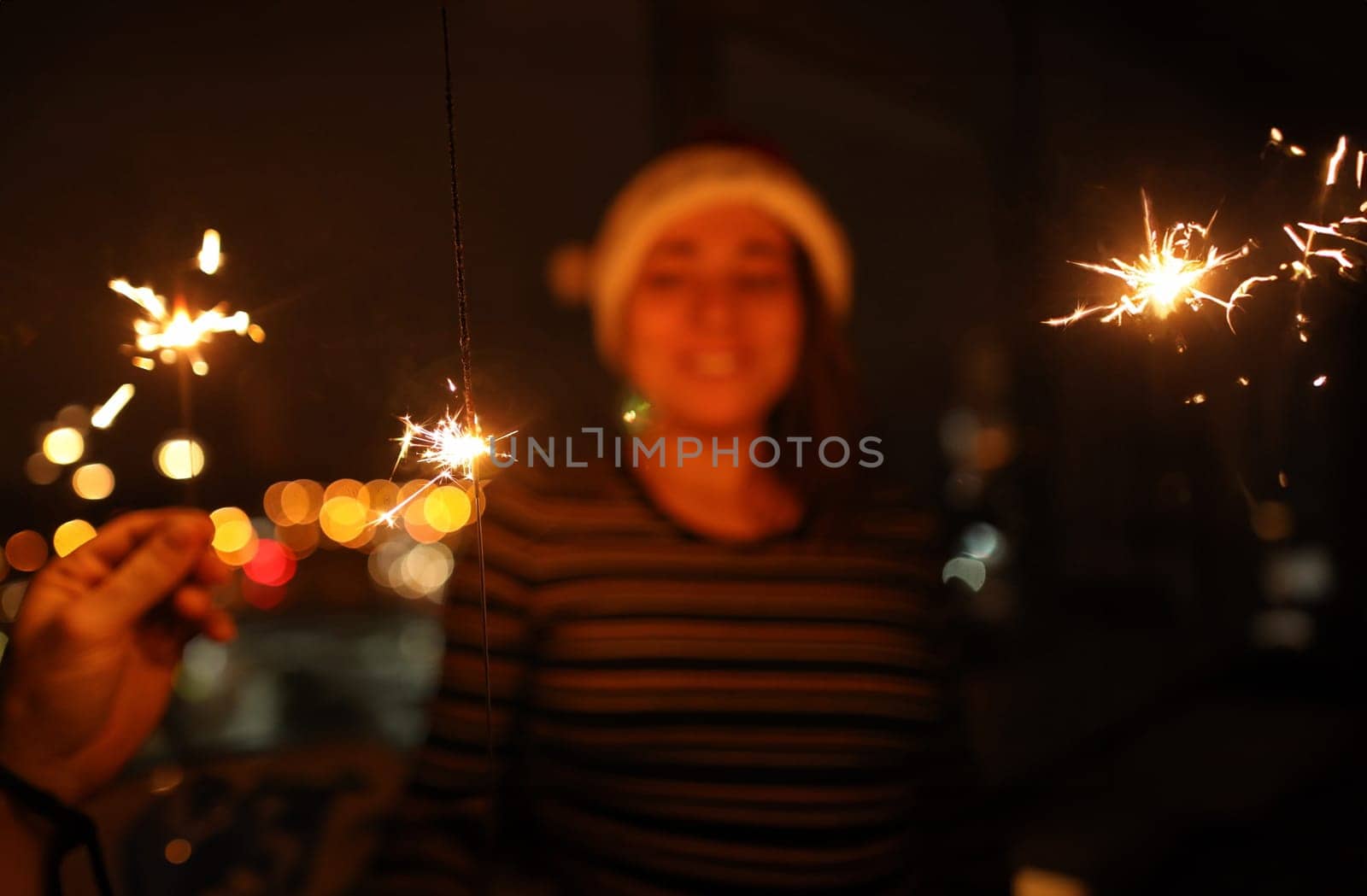 Sparkler in hand burn brightly. In background is woman with santa claus hat.