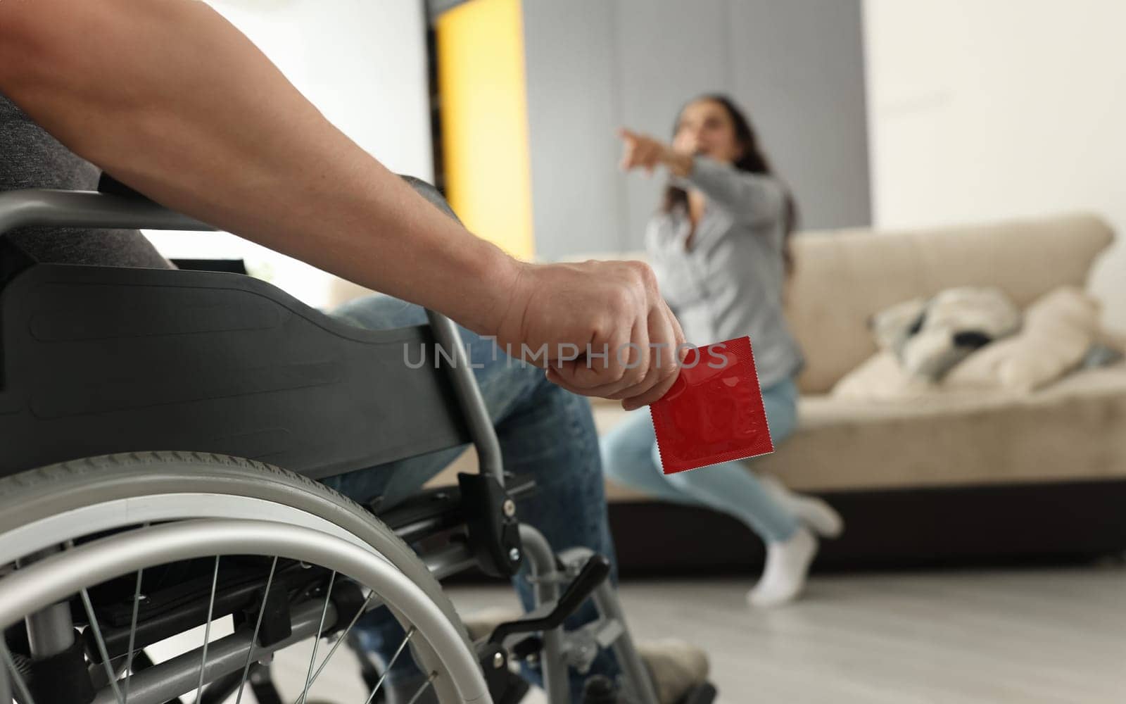 Woman sit on couch, point finger and laugh. Man in wheelchair hold red condom in his hand close up.