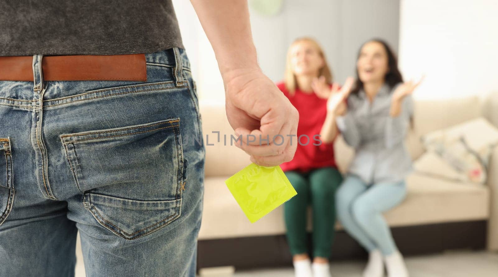 Two happy woman sit on couch and clap their hands. Man hold yellow condom in hand close up.