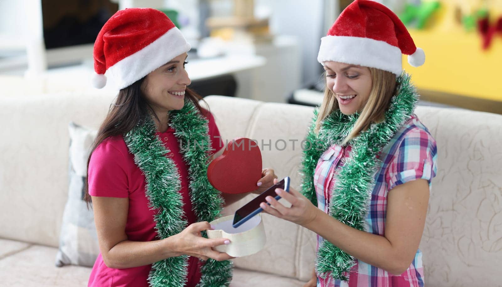 Beautiful woman in santa claus hat and tinsel around her neck give gift at form of heart. Surprised girlfriend open gift and take out phone.