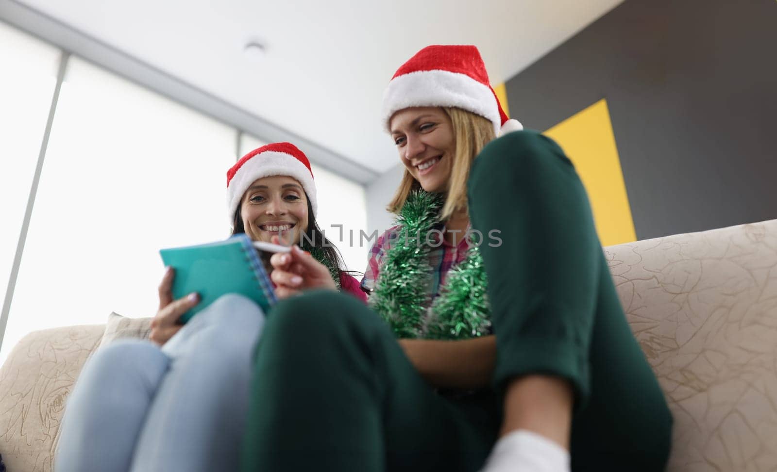 Two happy woman make up wishlist at home. Girlfriends sit on couch and discuss list in notebook.