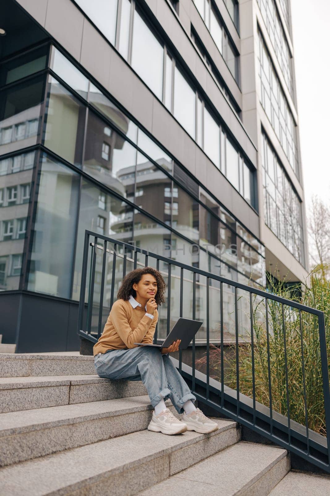Stylish female entrepreneur working on laptop sitting outside on modern building background