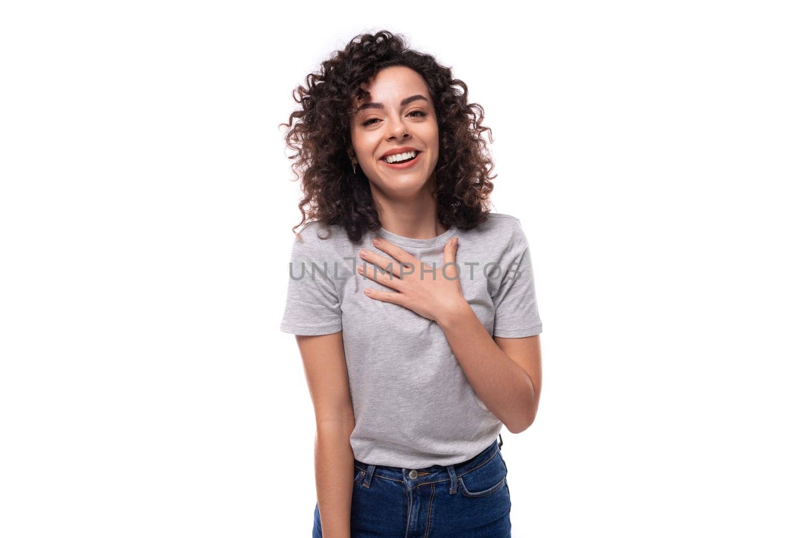 kind slender young caucasian woman with careless black curly hair is dressed in a gray t-shirt on a white background.