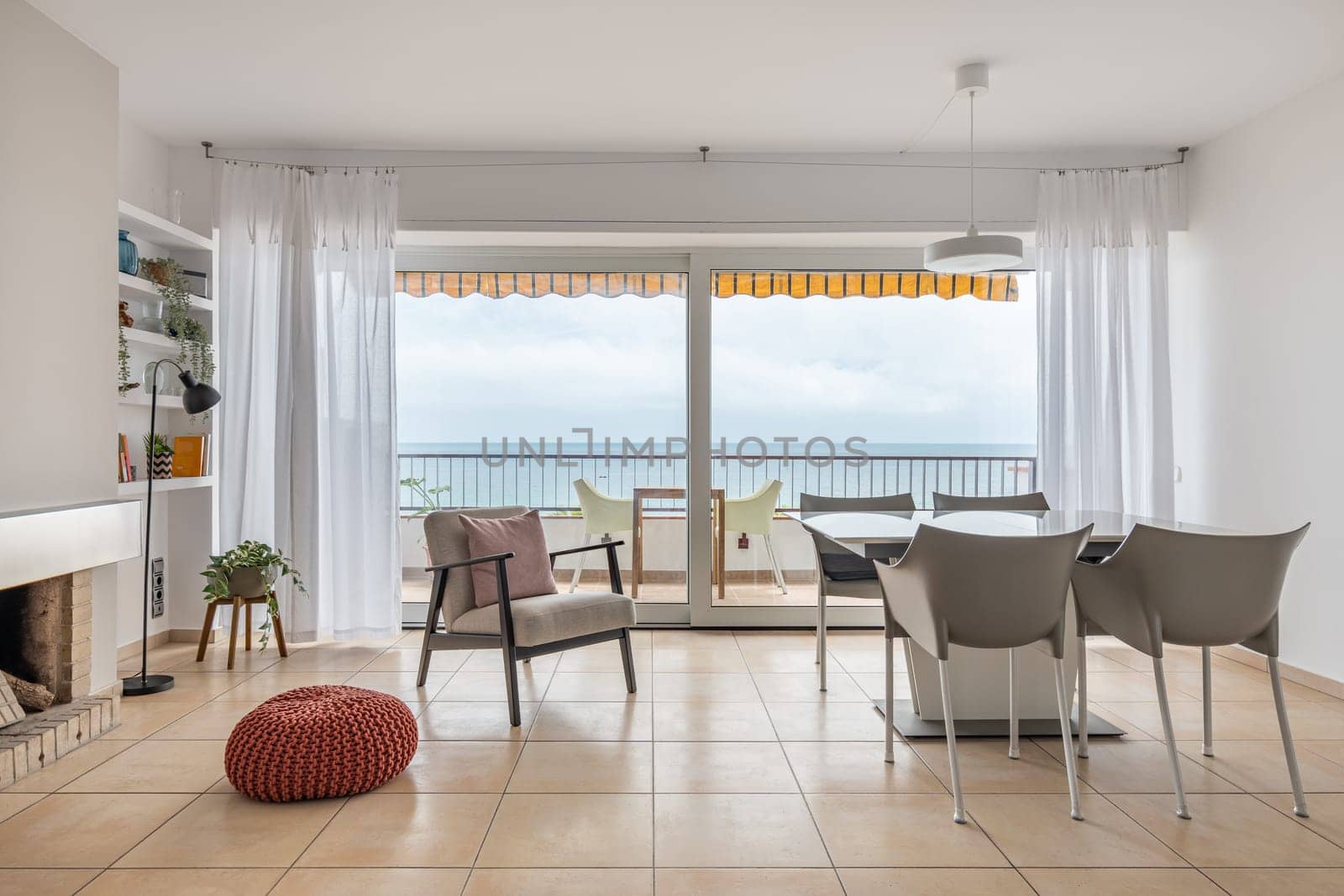 Dining table and soft armchair near terrace with panoramic window in living room facing ocean. Place to rest in hotel room. Minimalist interior