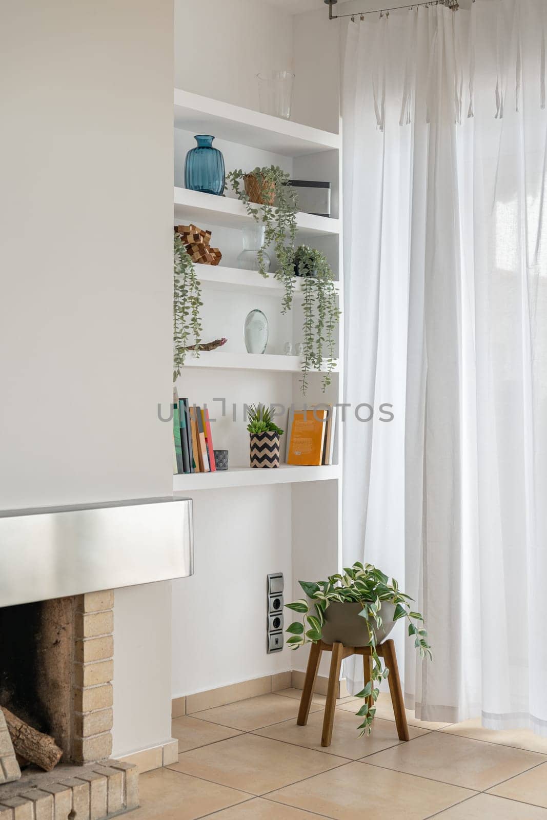 Shelving unit with pot plants and decorative details in living room. Bookshelves by window curtain in renovated apartment. Interior design