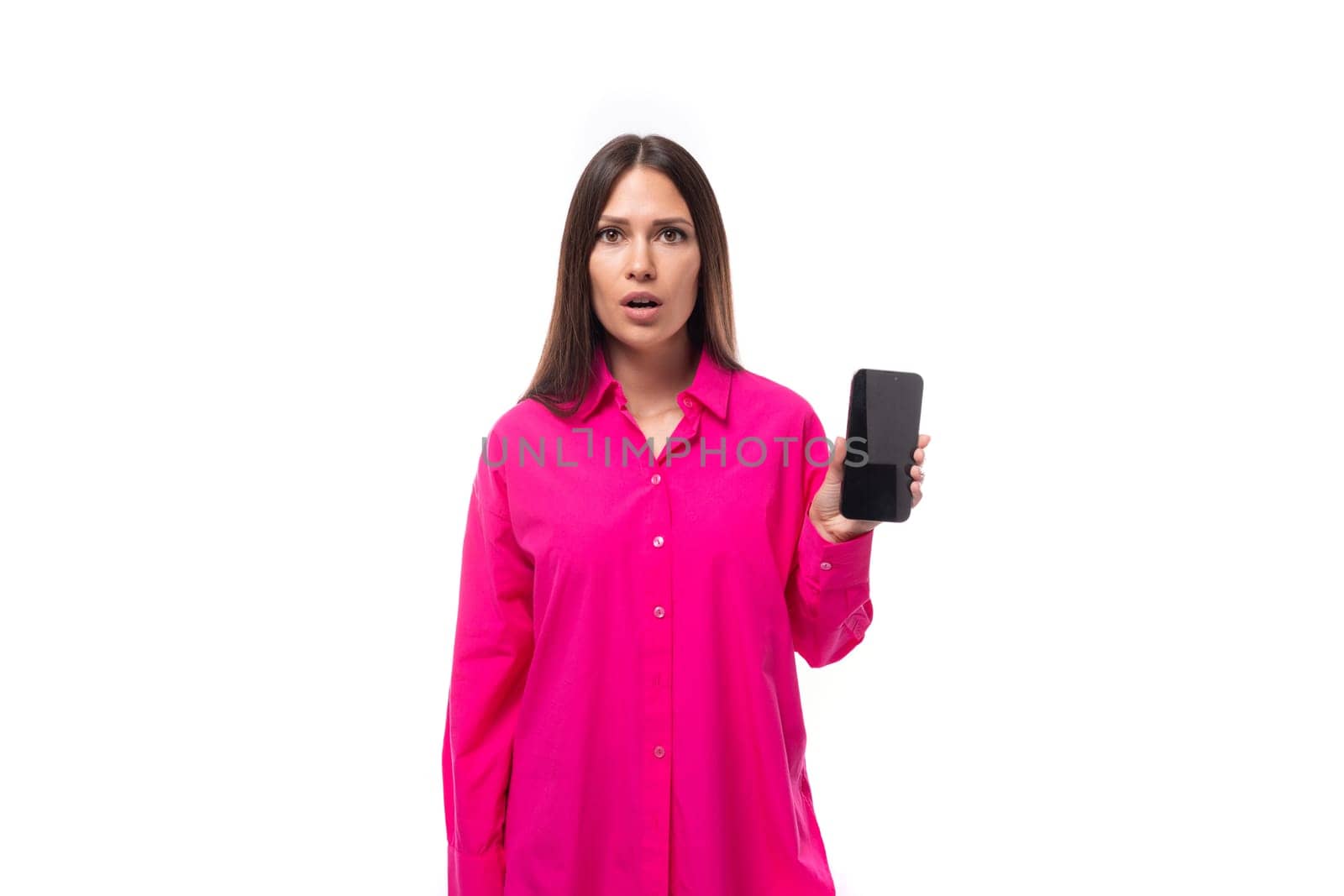 stylish caucasian woman with black straight hair dressed in a crimson shirt shows the smartphone screen in surprise.