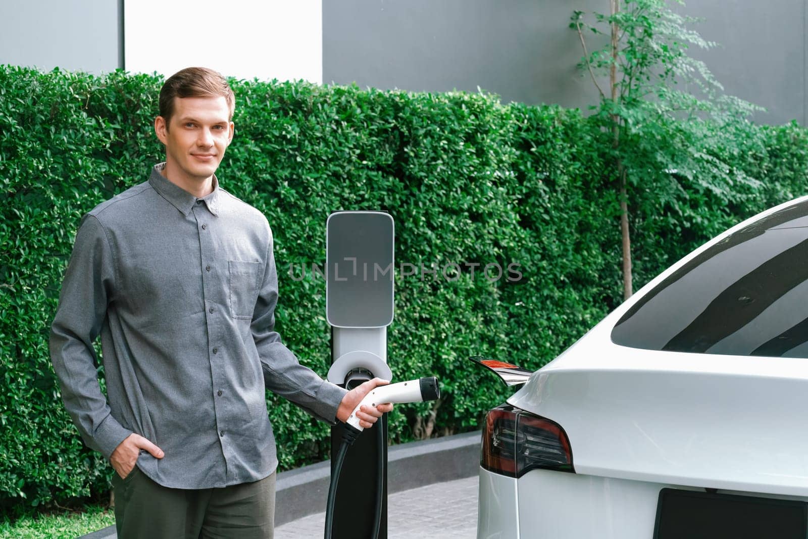 Young man travel with EV electric car charging in green sustainable city outdoor garden in summer shows urban sustainability lifestyle by green clean rechargeable energy of electric vehicle innards