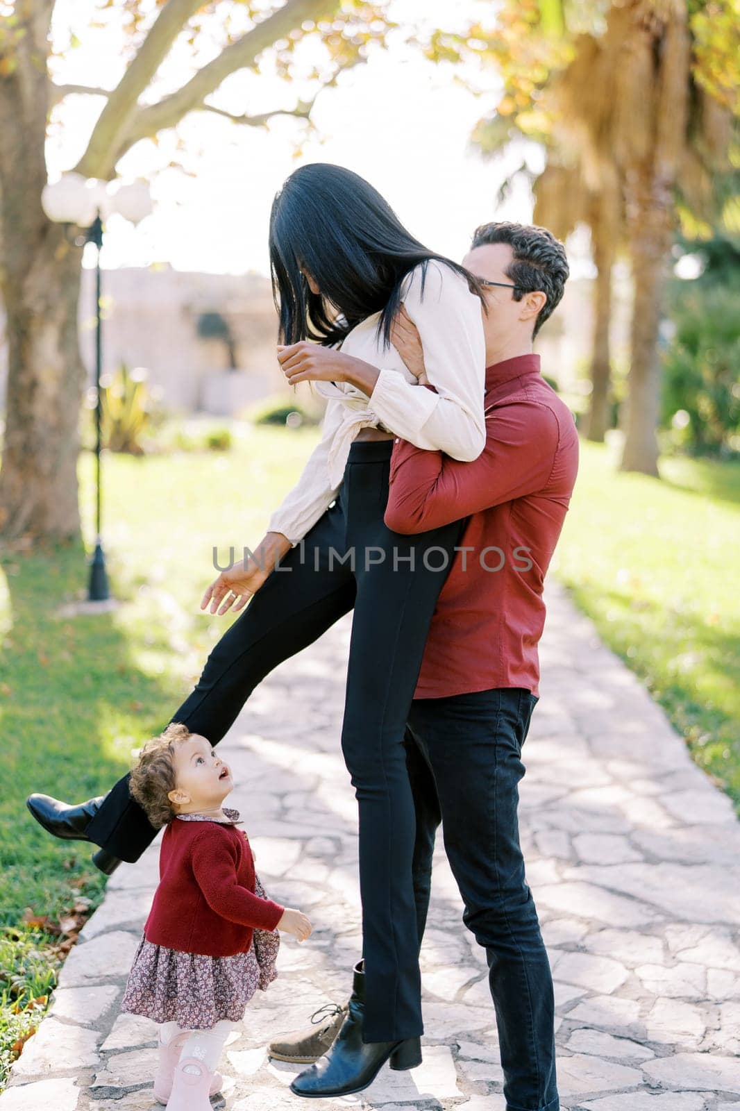 Little girl stands and looks with surprise at her father, who raises her mother in his arms by Nadtochiy