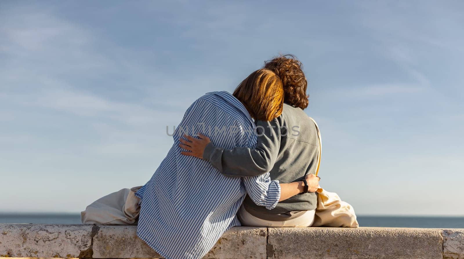Rear view of young couple is hugging while sitting on bench and enjoys beautiful ocean view by Yaroslav_astakhov