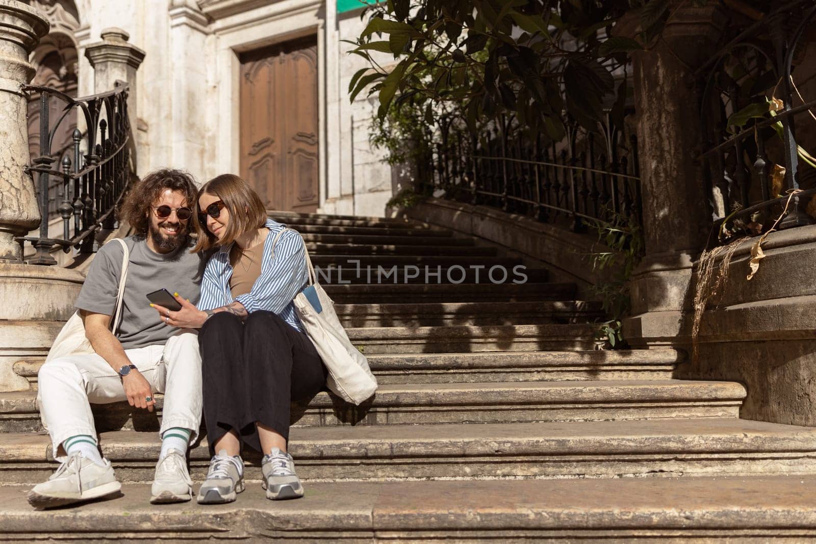 Beautiful couple in love sitting stairs on old city street and looking on phone. High quality photo