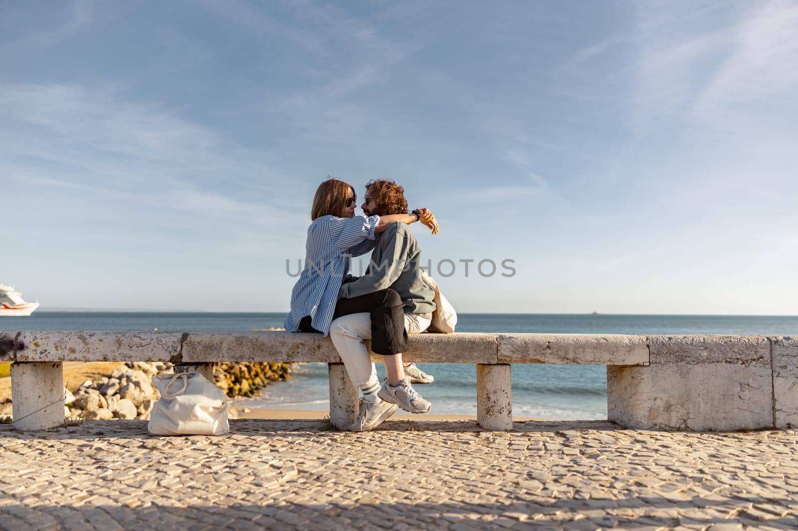 Couple in love is hugging and look into each other's eyes while sitting bench on beach near ocean by Yaroslav_astakhov