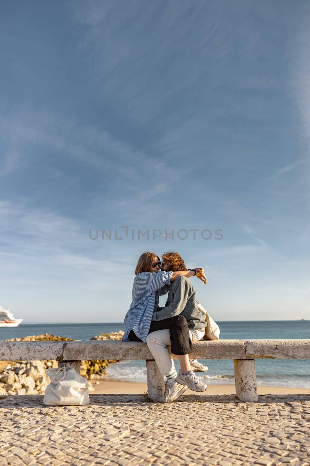 Couple in love is hugging and kissing while sitting bench on beach near ocean by Yaroslav_astakhov