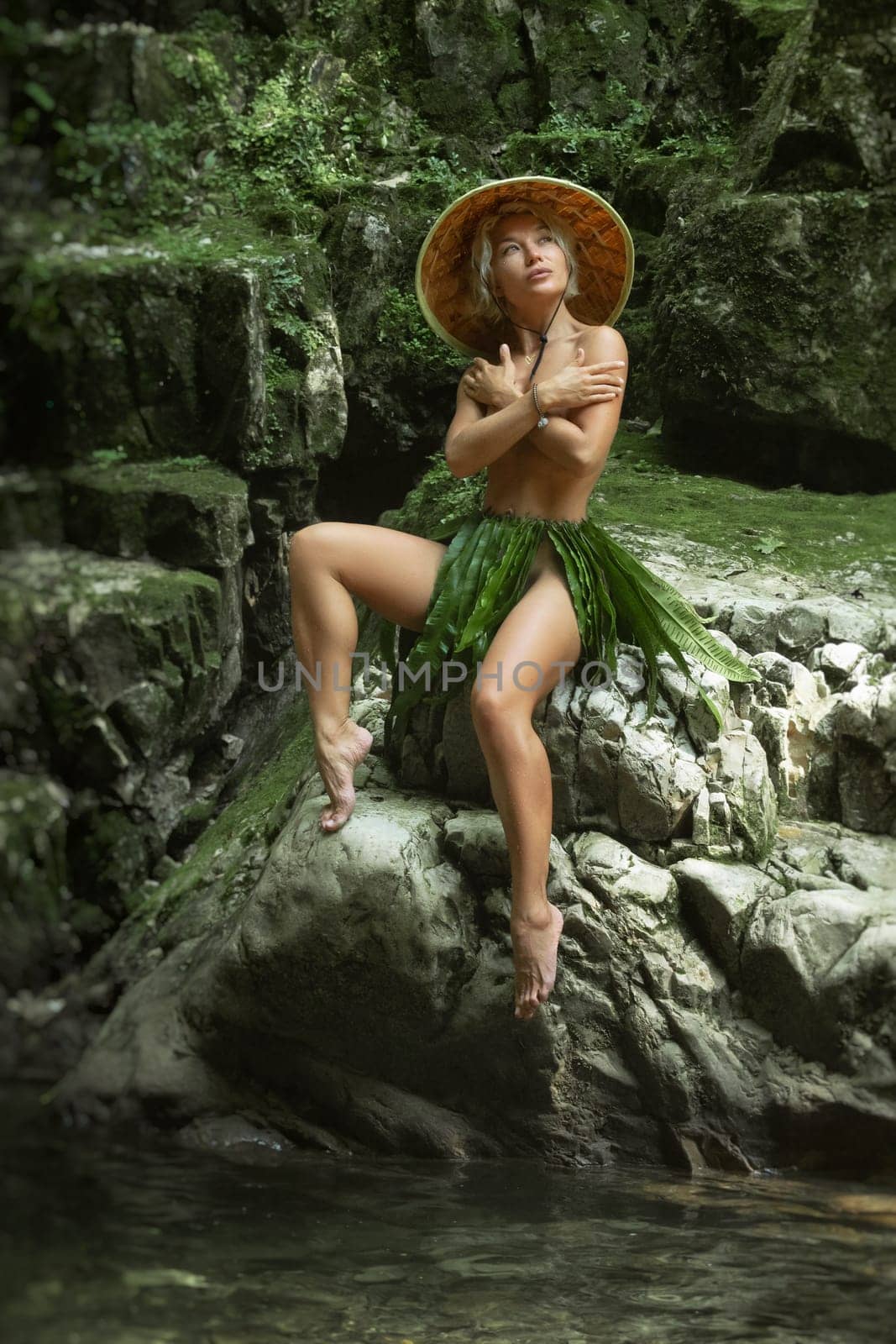 naked sexy girl in a triangular hat and a homemade cape made of leaves stands at the waterfall of a beautiful mountain river