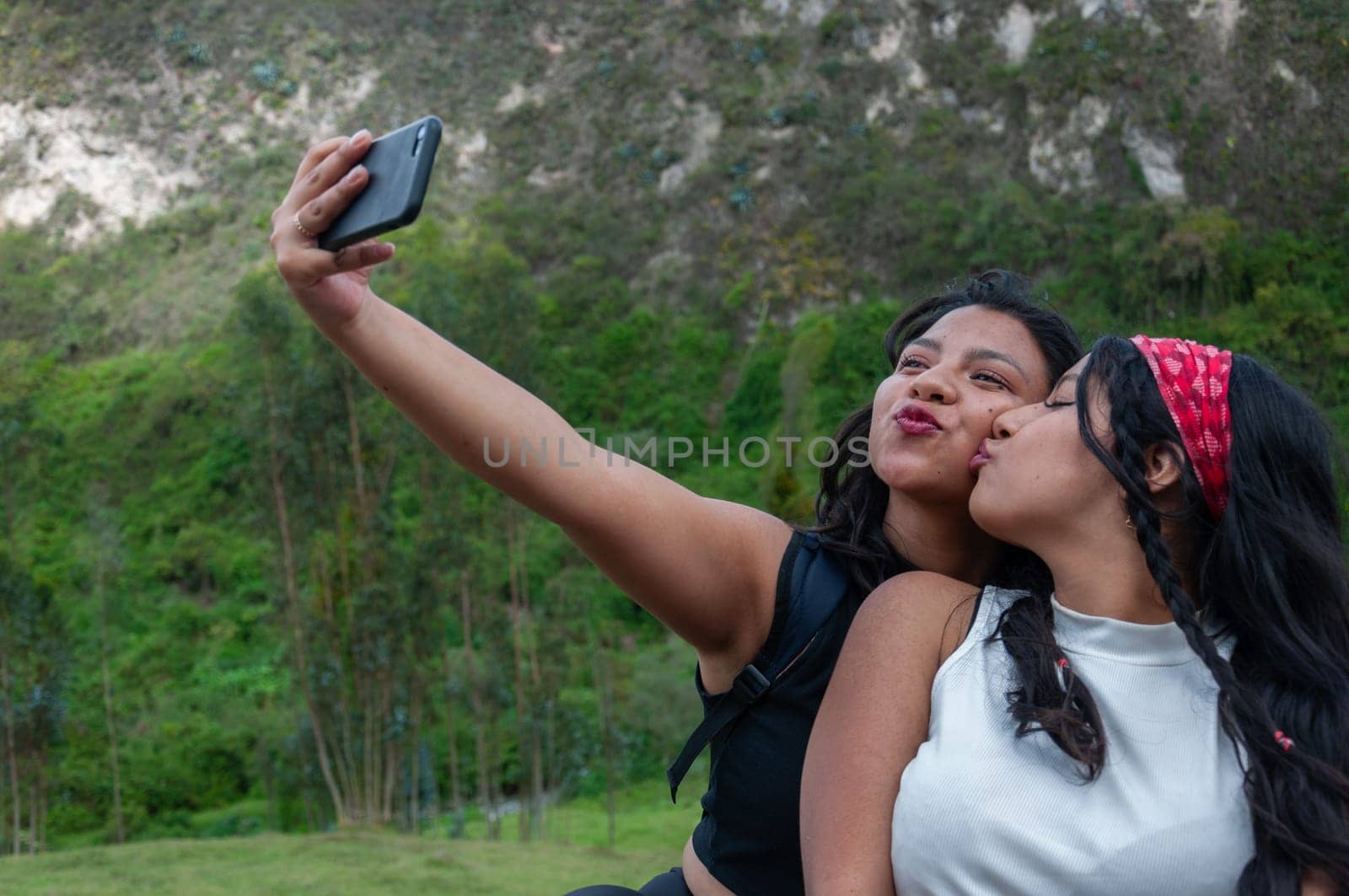 two friends taking a selfie at the top of a mountain, one looking at her cell phone and the other giving her friend a kiss. High quality photo