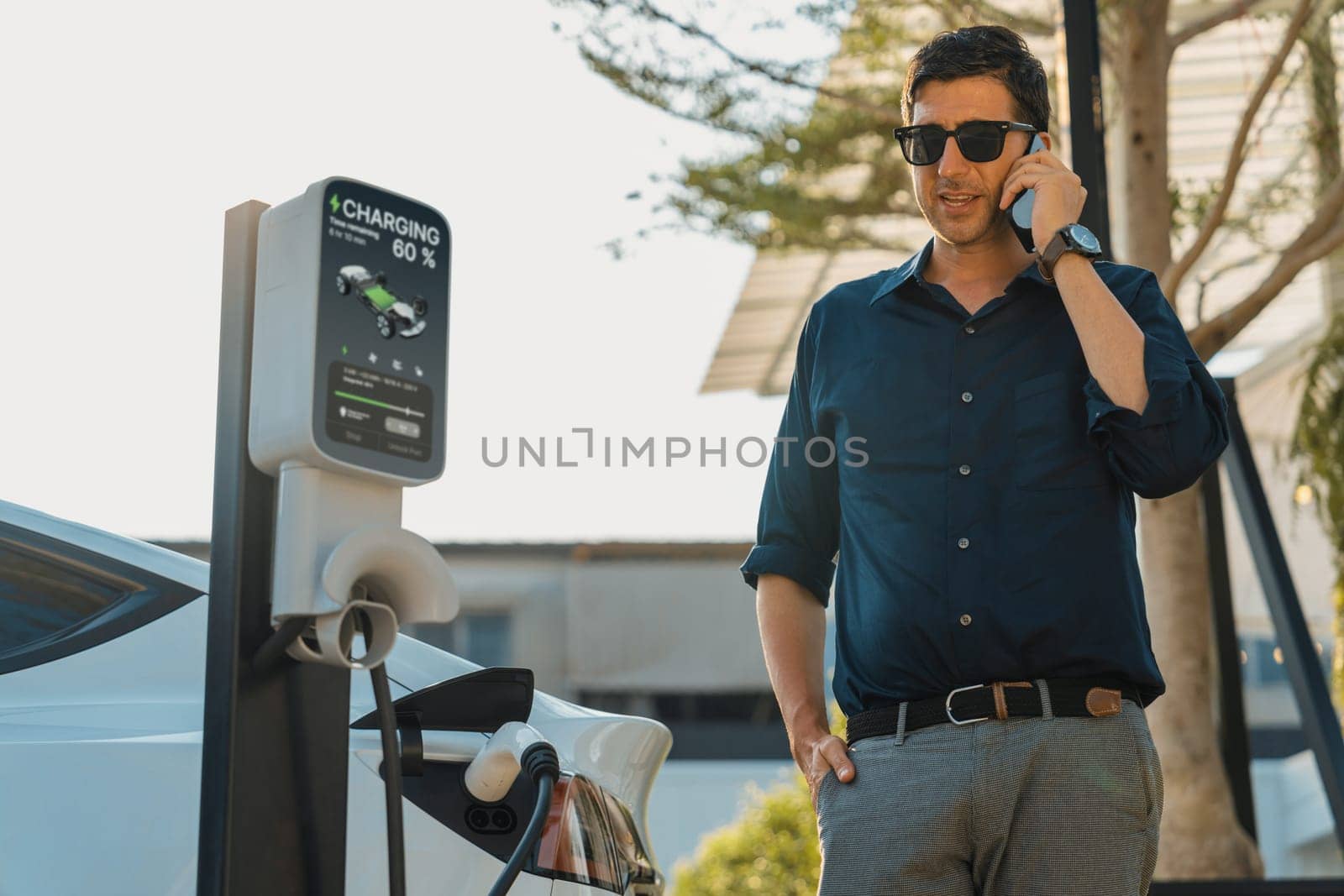 Young man recharge EV electric vehicle at green city park parking lot while talking on phone. Sustainable urban lifestyle for environment friendly EV car with battery charging station. Expedient